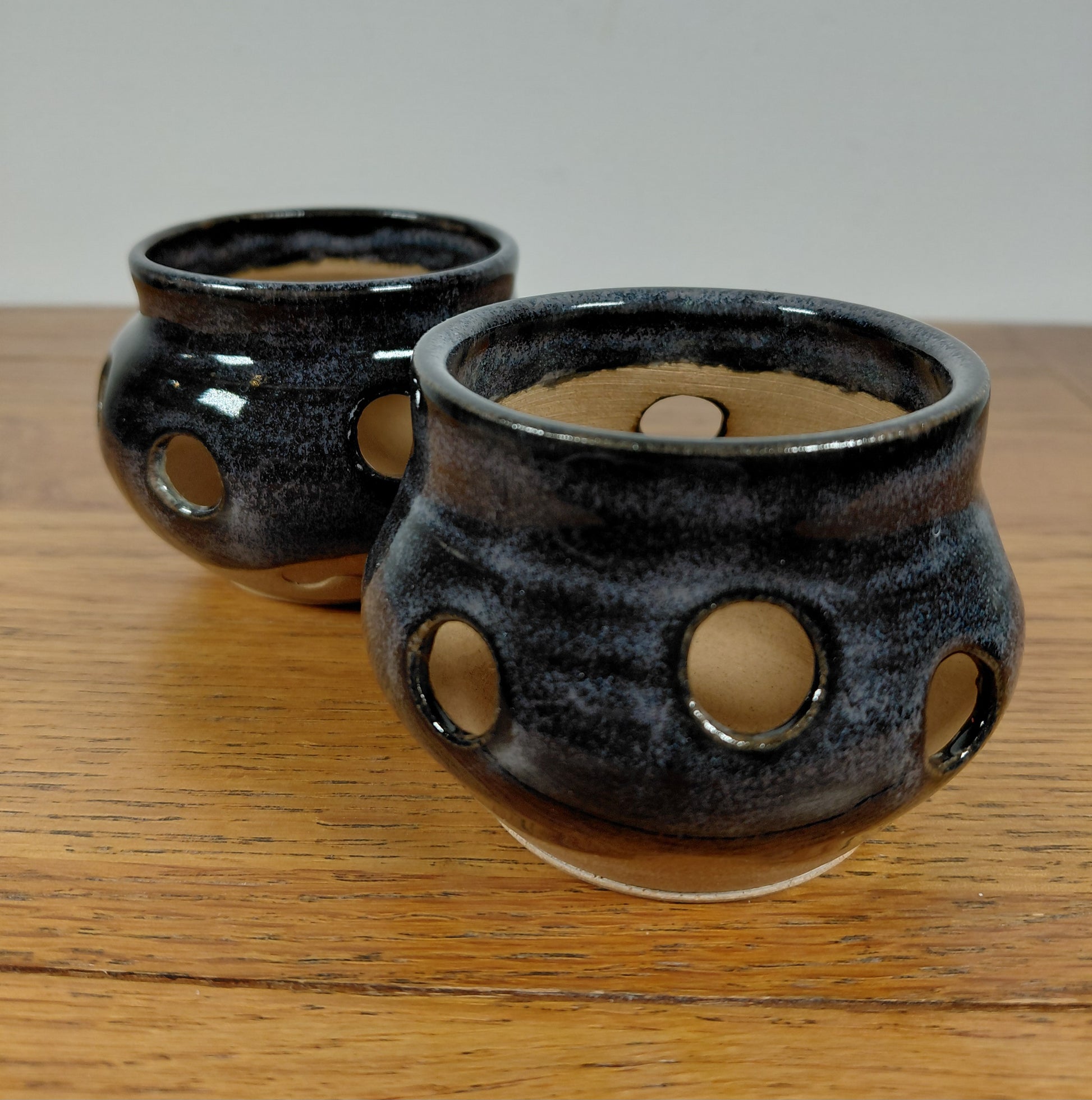 Two black ceramic pots with circular holes standing on a wooden surface