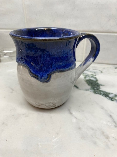 A handmade ceramic mug with a deep cobalt blue glazed top and handle, fading into a matte off-white glazed body, set on a white marble surface.