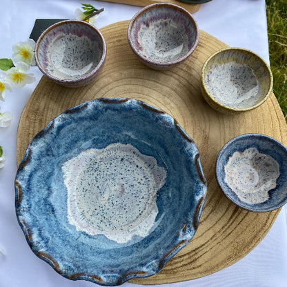 A set of handmade ceramic tableware, featuring a large scalloped blue plate with a white speckled center, four matching small bowls, and other plates, arranged on a wooden tray over a white tablecloth.